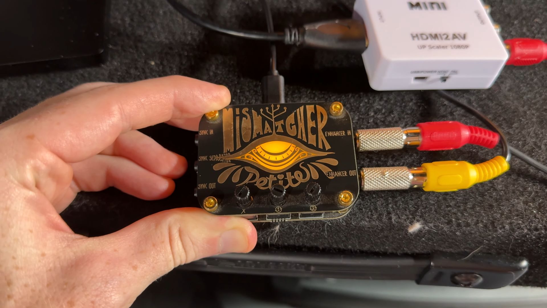 A hand holding a device labeled Mismatcher Petite with artistic design, connected to RCA cables and an HDMI to AV converter in the background.